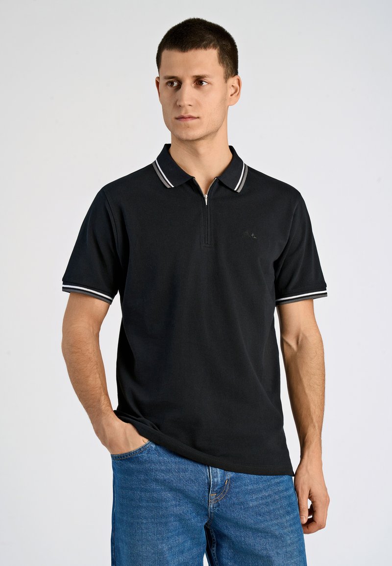 Svart pikétröja med dragkedjekrage och vita randiga detaljer på ärmarna, kombinerad med blå jeans. Slät tygstruktur.