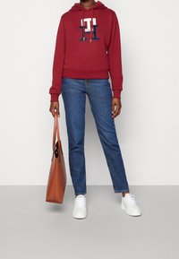 Sudadera roja con logo blanco y azul marino, combinada con jeans azules y zapatillas blancas. Lleva una bolsa de mano marrón en la mano izquierda.