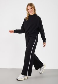 Czarna bluza z kapturem o luźnym kroju i ściągaczami, w połączeniu z czarnymi spodniami z białymi paskami po bokach i rozszerzanymi nogawkami. Sportowe sneakersy.