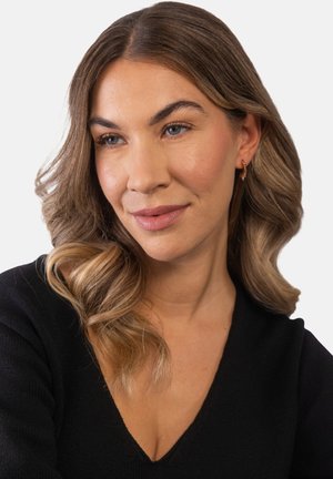 Frau mit welligem hellbraunem Haar, die goldene Creolen und ein schwarzes V-Ausschnitt-Oberteil trägt und leicht seitlich mit neutralem Gesichtsausdruck schaut.