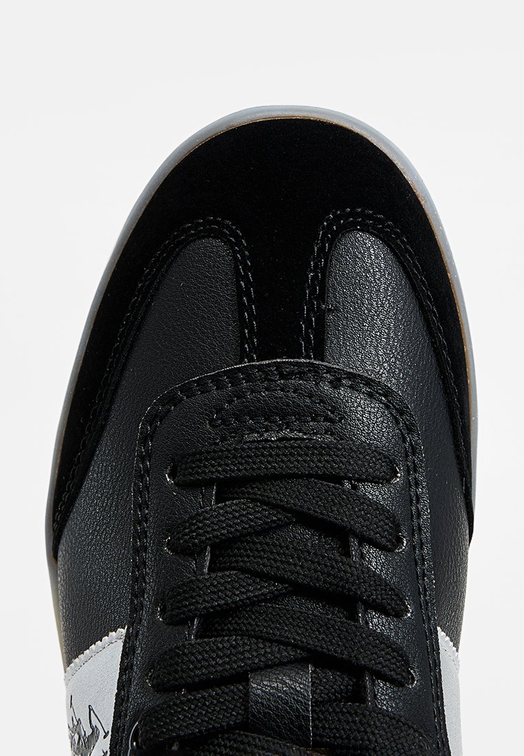 Zapatilla negra con una combinación de cuero suave y tejido texturizado, con cordones negros, un panel lateral blanco y una suela de goma.