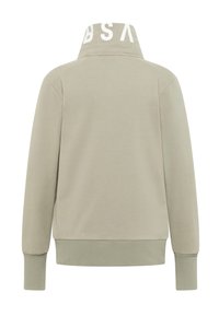 Svetlosivá mikina s vysokým golierom, žíhanými manžetami a lemom, s veľkým bielym logom na golieri. Vyrobená z mäkkej, hladkej látky.