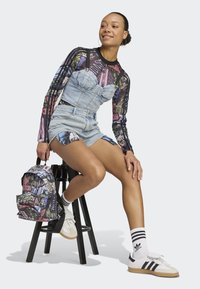 Denim overall shorts with a sweetheart neckline, paired with a long-sleeve graphic top featuring patterned details, white sneakers, and a patterned backpack.