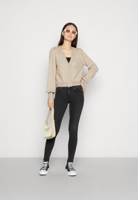 Cardigã de malha bege com decote em V e botões à frente, combinado com jeans pretos e ténis de cano alto azuis. O modelo segura uma mala creme.