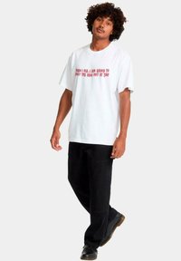 Hombre con cabello rizado vistiendo una camiseta blanca con texto rojo y pantalones negros, de pie con un pie ligeramente elevado sobre un fondo blanco.