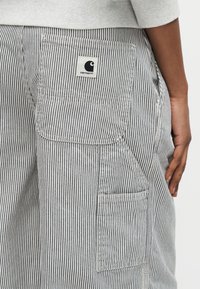 Οπίσθια όψη ατόμου που φοράει παντελόνι με κάθετες γκρι και λευκές ρίγες, με πολλαπλές τσέπες και ένα μπαλώμα λογότυπο Carhartt σε μία από τις τσέπες.