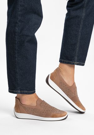Person trägt braune Slip-On-Sneaker mit geometrischen Mustern und weißen Sohlen, kombiniert mit dunklen Denim-Jeans vor einem einfarbigen Hintergrund.