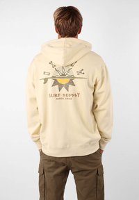 Haut à capuche beige clair avec un imprimé graphique présentant des éléments liés au soleil et au surf au dos. Coupe classique, texture douce et capuche avec cordon de serrage.