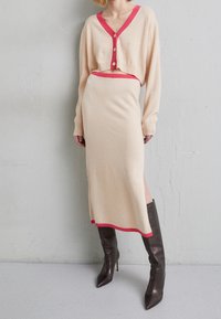 Beige neulepuku, jossa on lyhyt neulejakku vaaleanpunaisilla yksityiskohdilla ja hame sivuhalkiolla, yhdistettynä ruskeisiin korkokenkiin.