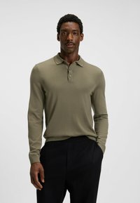 Mann in olivgrünem Langarm-Polohemd mit Knopfkragen und schwarzen Hosen, der mit einer Hand in der Tasche vor einem schlichten Hintergrund steht.