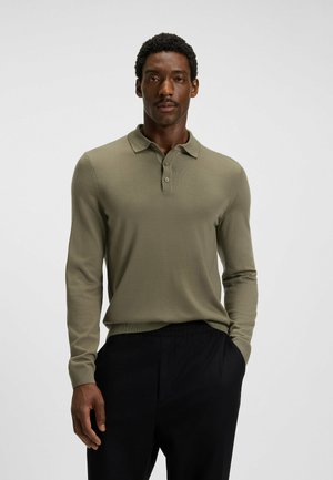 Mann in olivgrünem Langarm-Polohemd mit Knopfkragen und schwarzen Hosen, der mit einer Hand in der Tasche vor einem schlichten Hintergrund steht.