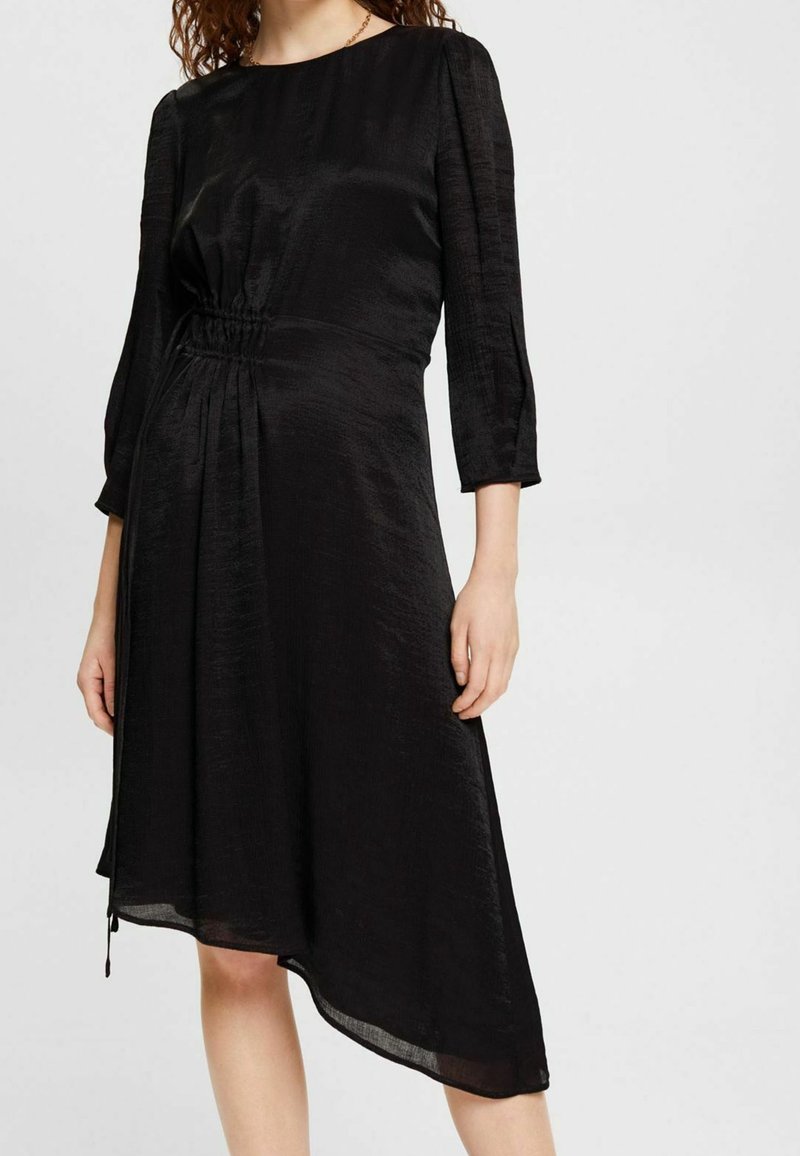 Femme portant une robe noire à ourlet asymétrique avec manches longues et détail froncé à la taille, montrée des épaules aux genoux.