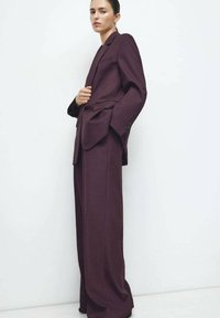 Costume sur mesure bordeaux avec un blazer structuré et un pantalon à jambes larges, confectionné dans un tissu texturé, avec des détails subtils de boutons.