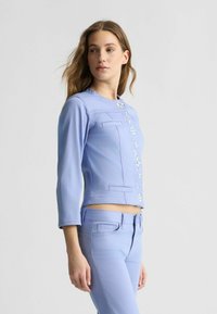 Hellblaues Jeansjackett mit Dreiviertelärmeln, Knopfverschluss und frontalen Verzierungen. Kombiniert mit passenden hellblauen Hosen.