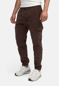Pantalones jogger de pana marrón con cinturilla elástica, bolsillos laterales y puños ceñidos, combinados con zapatillas deportivas gris claro.