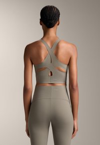 Olivno zelena športna crop majica s križnim hrbtom in izrezanimi detajli. Gladka tekstura z blagovno znamko elastičnim pasom na dnu.