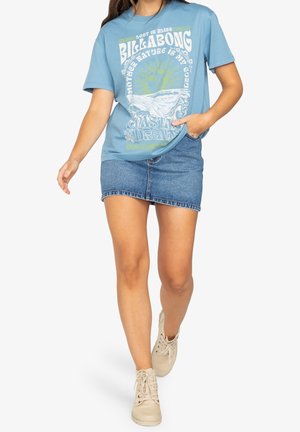 Kvinde går iført lyseblå grafisk Billabong t-shirt, denim mininederdel og beige snørestøvler på hvid baggrund.