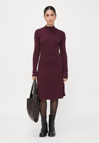 Bordeaux ribbed knit dress with long sleeves and a mock neck. Paired with a brown tote bag and black ankle boots, accentuated by fitted design.
