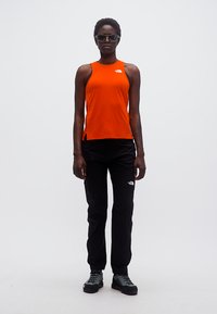 Άτομο που φοράει γυαλιά ηλίου, φωτεινή πορτοκαλί αμάνικη μπλούζα, μαύρο παντελόνι και γκρι παπούτσια πεζοπορίας, στέκεται μπροστά σε απλό λευκό φόντο.