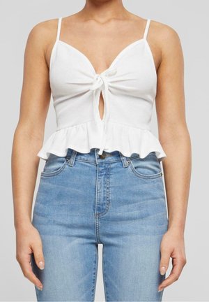 Femme portant un haut court blanc sans manches avec un nœud à l'avant et un ourlet à volants, ainsi qu'un jean en denim bleu clair taille haute, sur un fond gris.