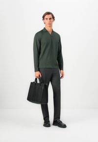 Man wearing dark green long-sleeve shirt, pinstripe trousers, black loafers, holding a large black leather tote bag, standing against a white background.