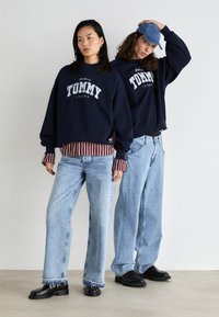 Sudadera marino con el logo "TOMMY JEANS" y dobladillo a rayas, combinada con jeans de color azul claro y zapatos negros. Vestimenta casual con un corte relajado.