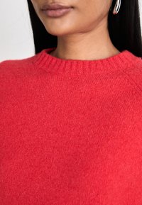 Czerwony sweter z dzianiny ze ściągaczem przy okrągłym dekolcie i gładką teksturą. Widok z bliska podkreśla detale szwów i jednolity kolor.