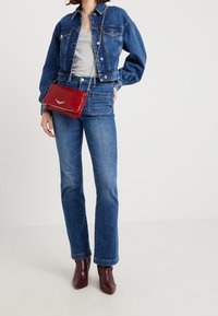 Mujer con chaqueta de mezclilla azul y jeans, camiseta gris, botas de tobillo burdeos, y llevando un bolso cruzado rojo con cadena plateada.