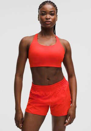 Atletische vrouw met gevlochten haar staat zelfverzekerd in bijpassende felrode sportbeha en shorts tegen een effen lichte achtergrond.