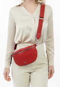 Borsa a tracolla in pelle rossa con forma curva, chiusura con zip argentata, tracolla in tessuto regolabile e un discreto dettaglio di branding nella parte anteriore.