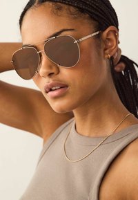 Aviators met bruine lenzen en een dunne, zilveren metalen rand. Model draagt een tan kleurige mouwloze top en een eenvoudige gouden ketting.