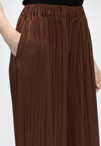Personne portant une jupe plissée marron avec une taille élastique et une poche latérale avec la main insérée, sur fond blanc.