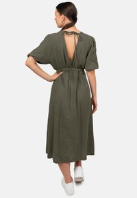 Olijfgroene, gestructureerde maxi-jurk met een strik achter, korte pofmouwen en een open rug, gecombineerd met witte sneakers.