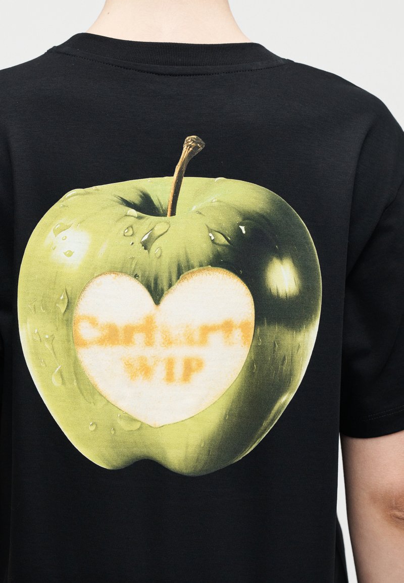 Grüner Apfel mit Wassertropfen und einem herzförmig geschälten Abschnitt, der den verblassten Schriftzug "Carhartt WIP" auf der Rückseite eines schwarzen T-Shirts zeigt.