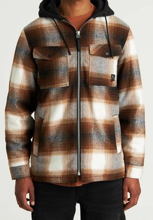 Uomo che indossa una giacca con cerniera a quadri marrone, neri e bianchi, con cappuccio nero e due tasche con patta e bottoni sul petto.