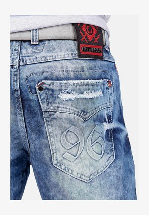Jeans en denim bleu délavé avec des détails usés, des coutures contrastantes et une étiquette en cuir texturé noir et rouge sur la taille.