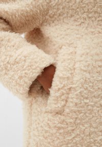 Manteau fluffy couleur crème avec un tissu texturé. Présente une grande poche sur le côté gauche. Surface douce et duveteuse avec des fibres visibles.