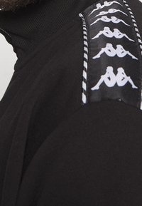 Felpa nera con collo alto, caratterizzata da un design del logo bianco lungo la spalla e una rifinitura a righe contrastanti sul lato.