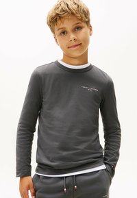 Blonder Junge, der einen dunkelgrauen Tommy Hilfiger Sweatshirt und passende Hose trägt, mit Händen in den Taschen, schaut in die Kamera vor einem weißen Hintergrund.