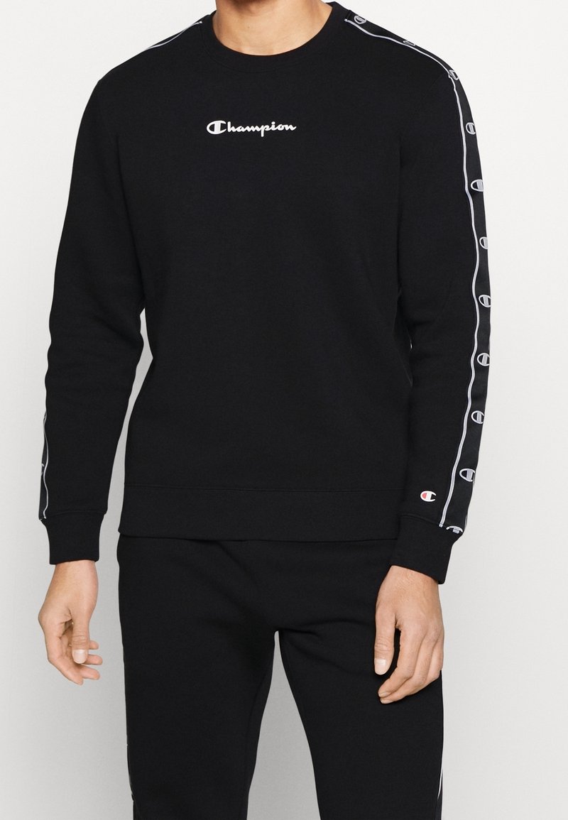 Homme portant un sweat-shirt noir Champion avec un logo blanc sur la poitrine et des logos répétés le long des manches, associé à un pantalon noir.