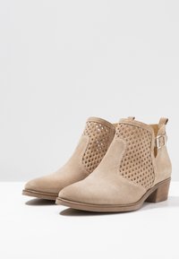 Beige nahka ankle boots, joissa on leikattu ristikkosuunnittelu, pyöreä kärki, matala pinottu korko ja sivulla oleva solkiyksityiskohta. Suora pinta.
