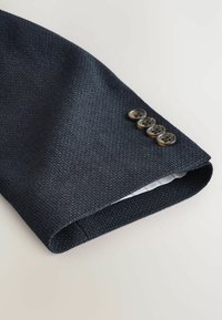 Navy strukturovaný oblek s rukávem zdobeným čtyřmi tmavými knoflíky; vnitřní látka má jemný kostkovaný vzor. Hladký povrch, moderní střih.