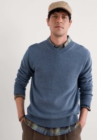 Pull en maille bleue avec un col en V, ourlet et poignets côtelés. Porté par-dessus une chemise à carreaux avec des nuances de bleu, vert et marron ; présente une casquette déstructurée.