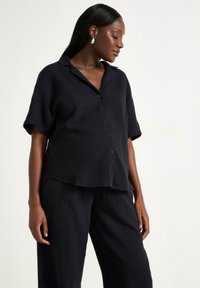 Camicia nera a maniche corte con collare, dal tessuto testurizzato, abbinata a pantaloni larghi coordinati. Tessuto morbido e stropicciato.