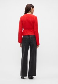 Červený svetr s dlouhými rukávy a přiléhavým pasem, spojený s černými kalhotami s širokými nohavicemi a černými botami. Měkká textura a jednoduchý design.