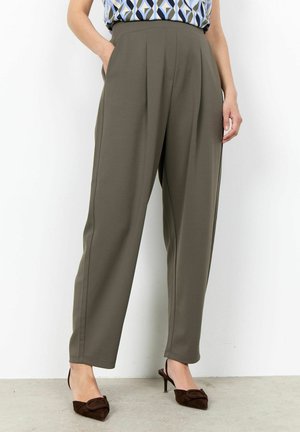 Persona che indossa pantaloni a pieghe a vita alta verde oliva con tasche laterali e tacchi a punta in camoscio marrone scuro su sfondo chiaro.