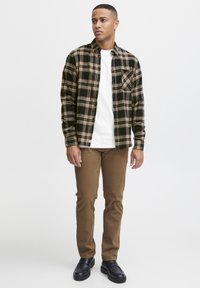 Koszula flanelowa w czarno-brązową kratę, noszona na białym T-shircie, w połączeniu z brązowymi spodniami slim fit i czarnymi butami.
