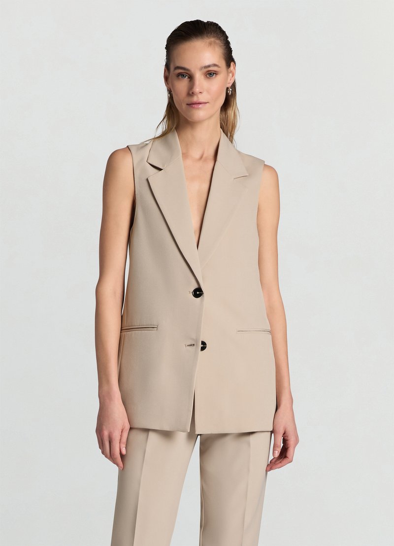 Blazer sans manches beige avec un col cranté, deux poches avant, des boutons noirs uniques et une texture de tissu lisse.