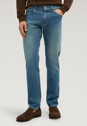 Hombre con vaqueros de corte recto azul medio, mocasines marrones y una camiseta marrón de manga larga, con una mano en el bolsillo.