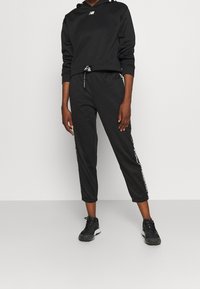 Svart huvtröja med New Balance-logga, ihop med svarta joggingsbyxor med vita sidodetaljer och sportskor.